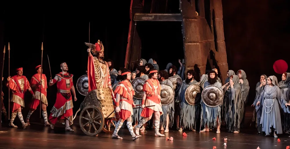 Escena de Nabucco: el coro con escudos y un carro sobre el escenario.
